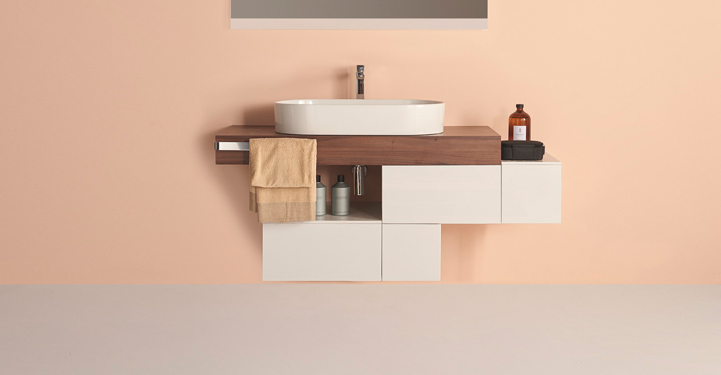 Photo de la collection de meuble Adapto mise en ambiance avec un plan beige, une étagère marron, une vasque et une robinetterie sur fond en brique gris