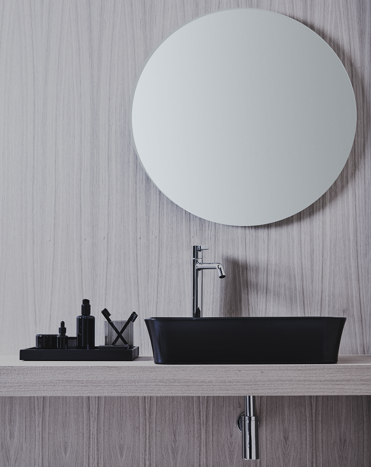 Photo d'une salle de bains montrant une vasque Ipalyss noire, une robinetterie Joy chrome sur un plan en bois surmontés d'un grand miroir rond